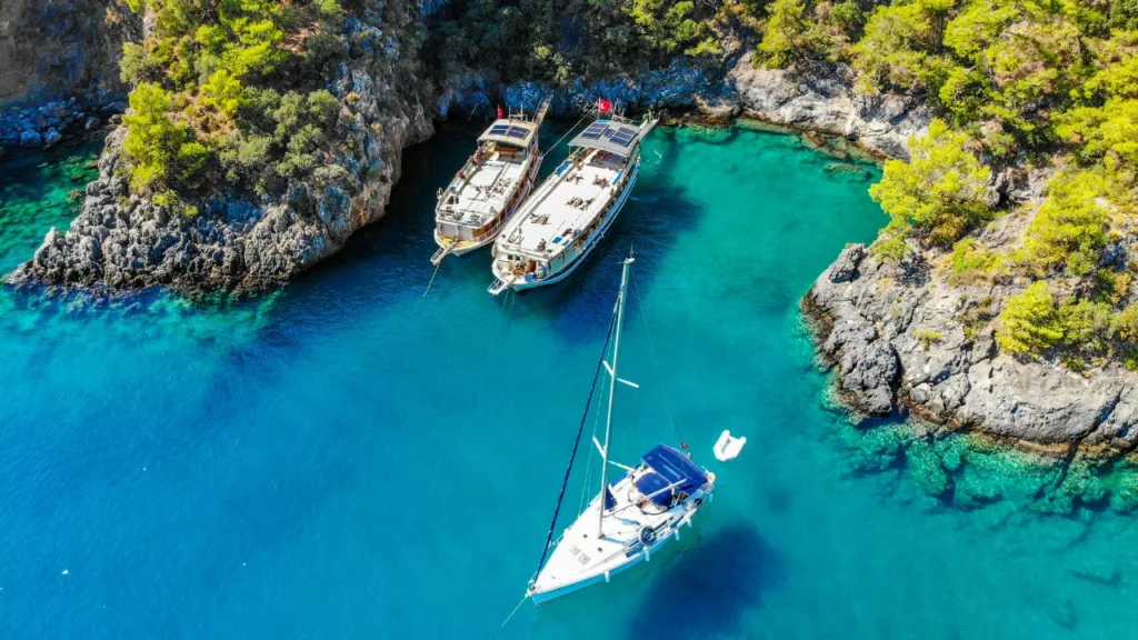 Paisaje rocoso lleno arboles con un lago hermoso de color azul, donde flotan 3 barcos esperando a turistas que contrataron a un agente de viaje.