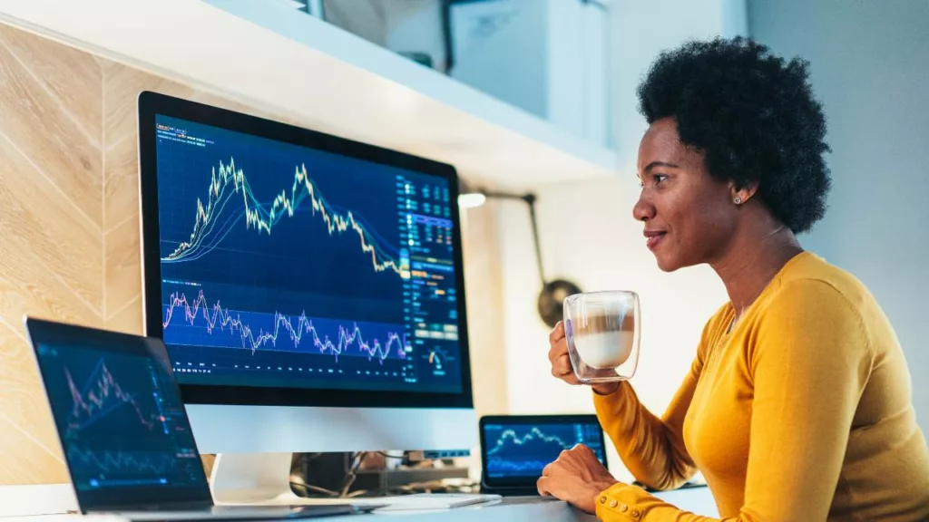 Corredora de bolsa de valores, revisando gráficos del mercado desde su computadora y tomando un café.