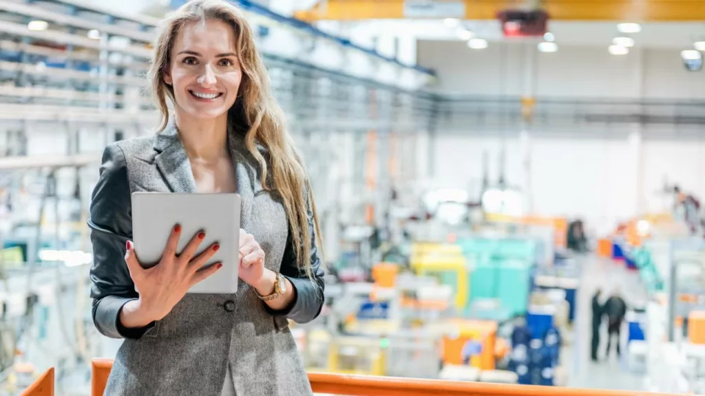 Administradora en una fabrica, con vestimenta profesional, sonriendo y sosteniendo su tablet de anotaciones.