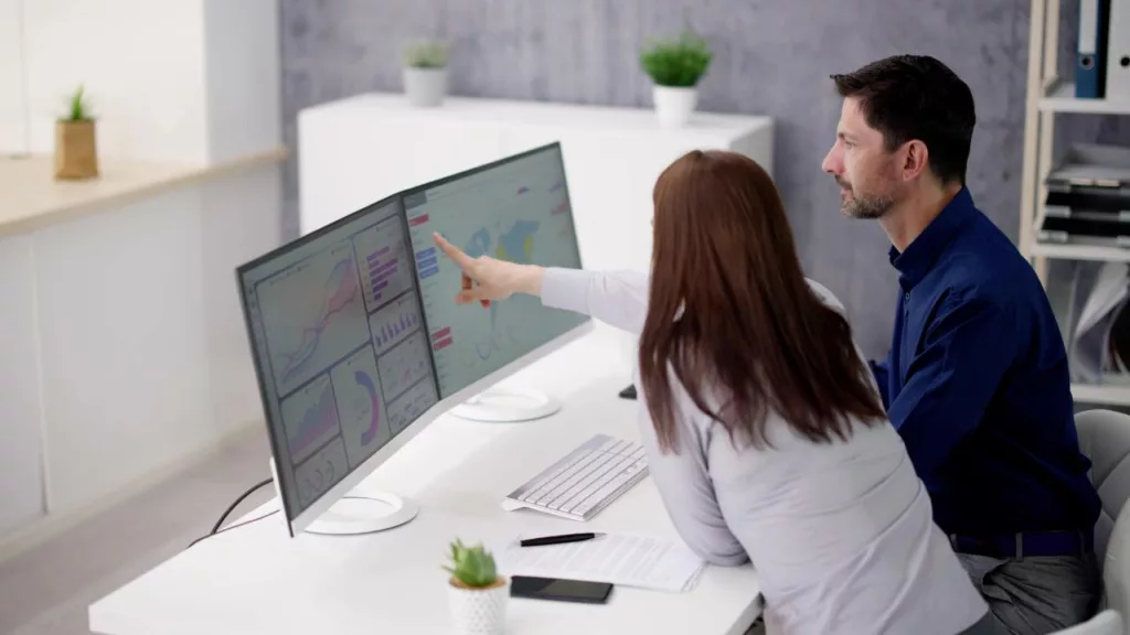 Mujer le señala un gráfico de análisis de la pantalla del computador de su compañero de trabajo.