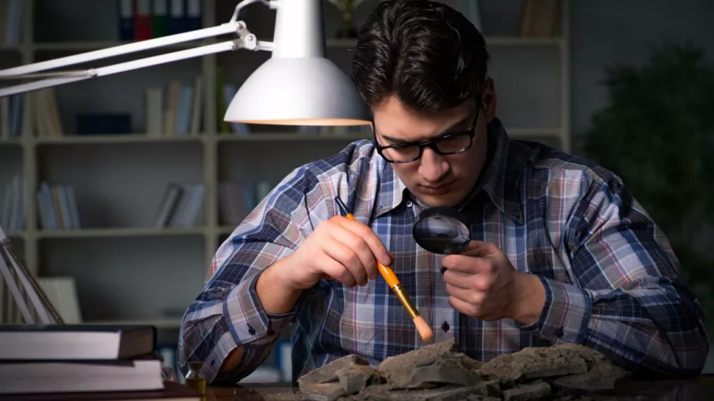 Antropólogo con una lupa y una brocha, estudiando una pieza de alguna civilización antigua.