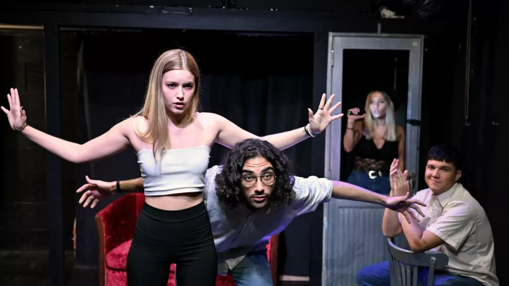 Actriz y actor, junto con otras 2 personas detrás, actuando una escena de una obra.