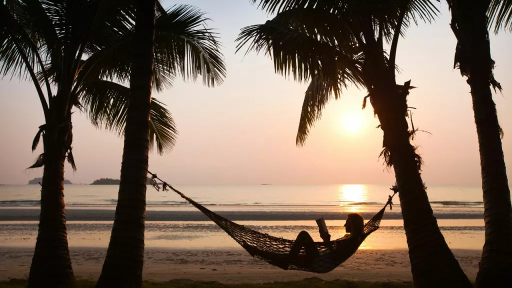 Playa con un atardecer, donde una mujer disfruta por un agente de viaje, el ambiente con una hamaca y un libro.
