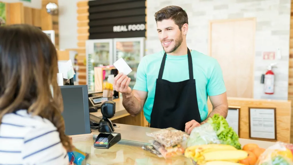 Hombre joven trabajando como cajero y atendiendo a una cliente, que desea facturar los alimentos.