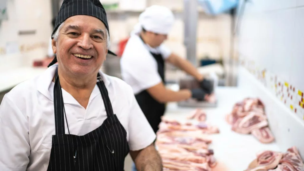 Jefe de carnicería trabajando y detrás un carnicero, cortando carne.