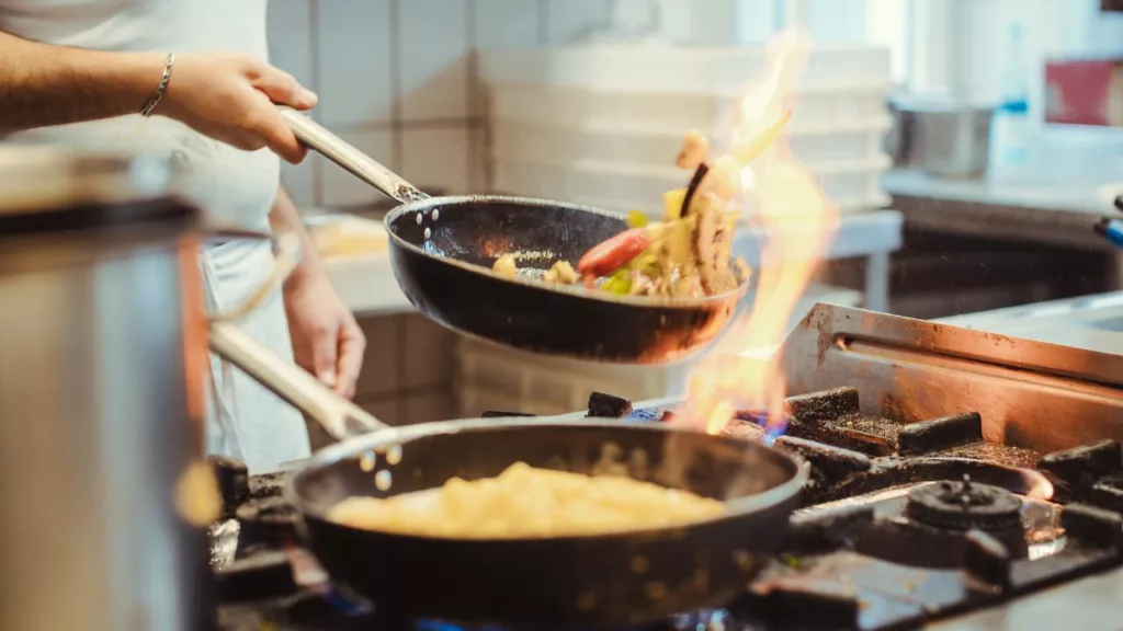 Cocinero usando dos sartenes para preparar alimentos en una estufa.