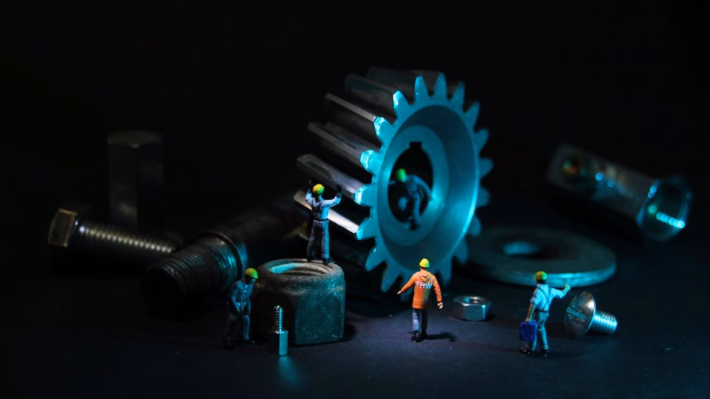 Fotografía artística macro de figuras en miniatura (trabajadores) interactuando con una gran rueda dentada (engranaje), tuercas, tornillos y otros componentes de Ingeniería Mecánica en un ambiente oscuro con luz azul y verde. La escena simboliza el trabajo en equipo y la escala de un proyecto realizado por un Ingeniero Mecánico.
